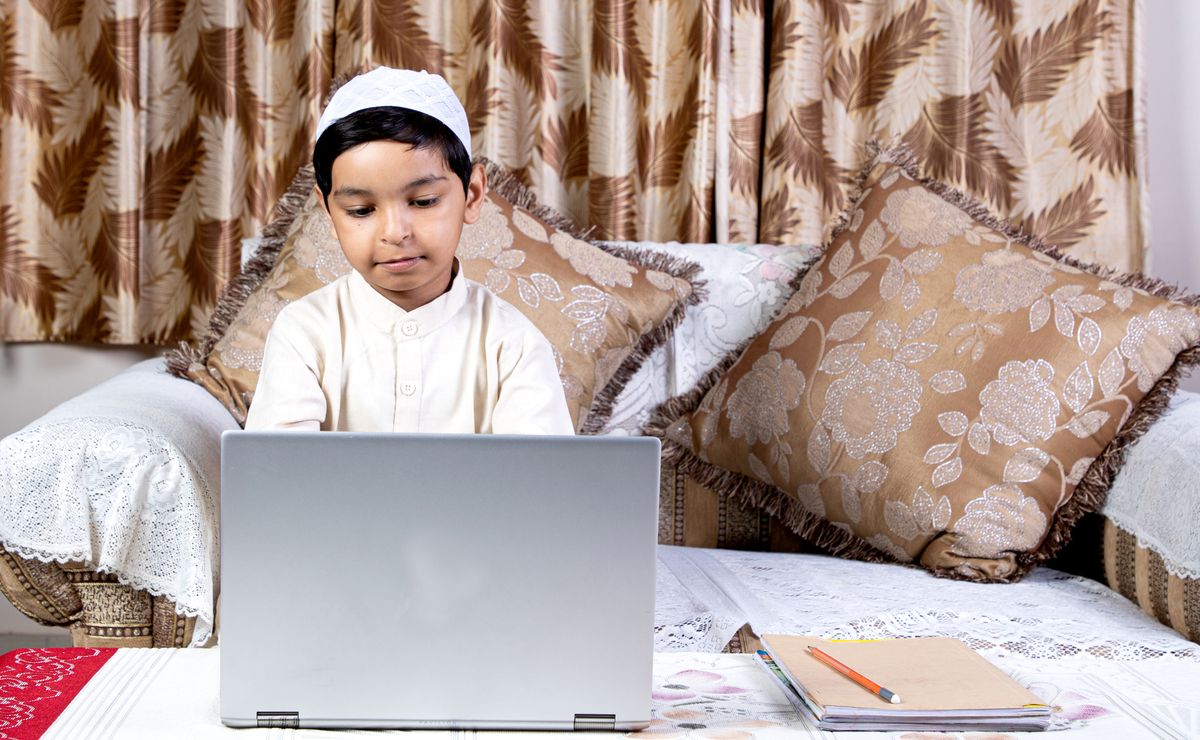 A boy in a white prayer cap and kurta works at a laptop on a sofa at home, a notebook and pencil beside him.