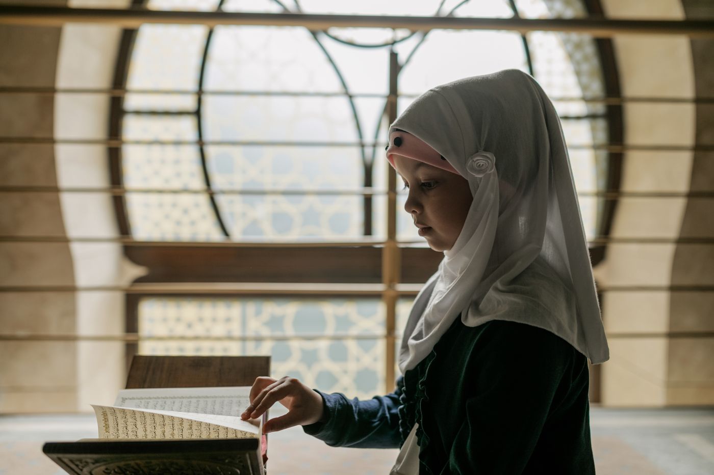 A young girl in a white headscarf reads the Qur'an on a wooden stand beside a sunlit window patterned with Islamic geometry.
