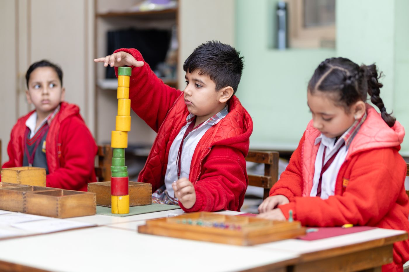 Three children in red school sweaters work with Montessori materials at a table; one stacks coloured wooden cylinders while another sorts beads.